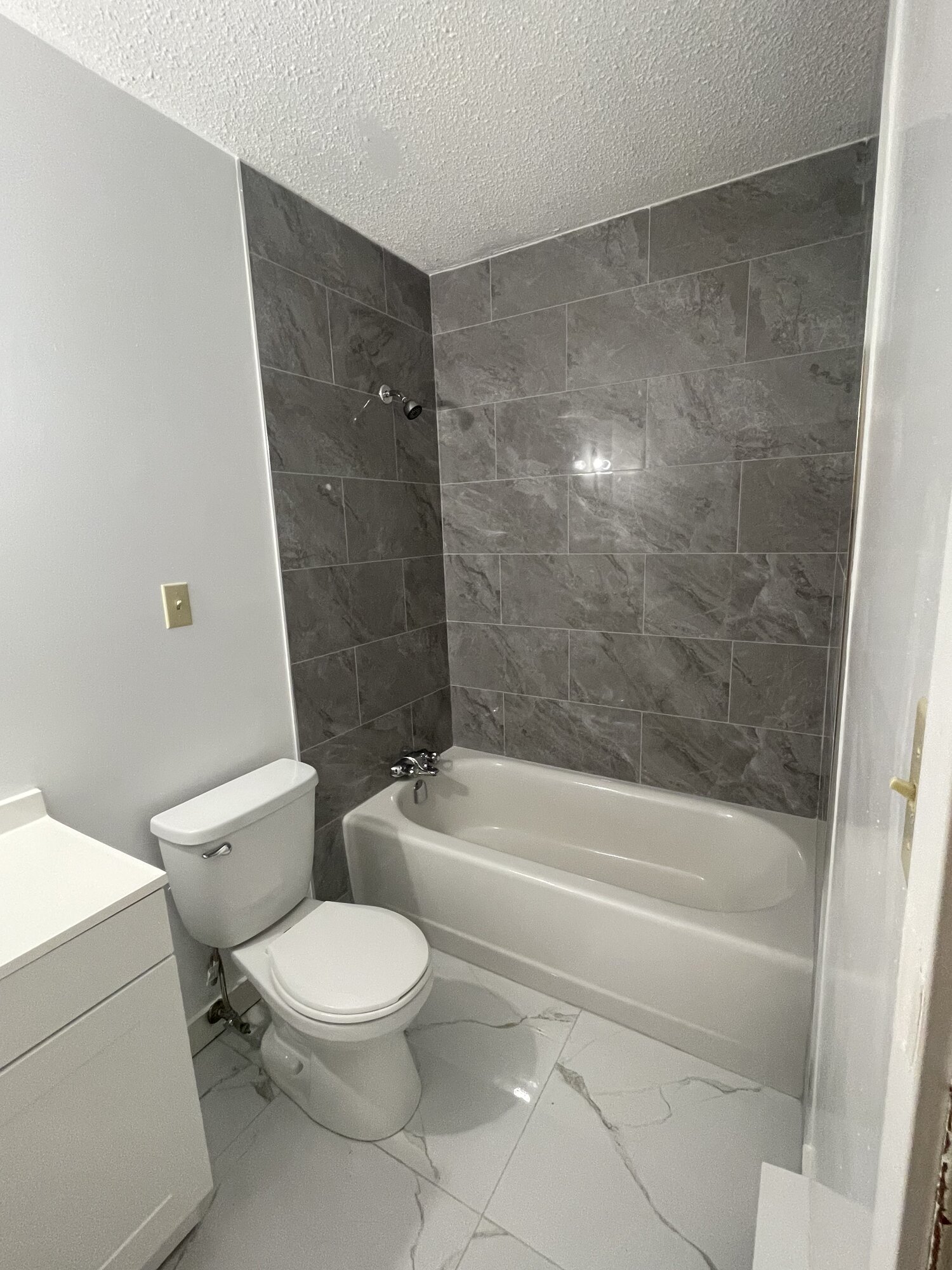 The same bathroom with large grey wall tile around the tub and a white marble look floor.