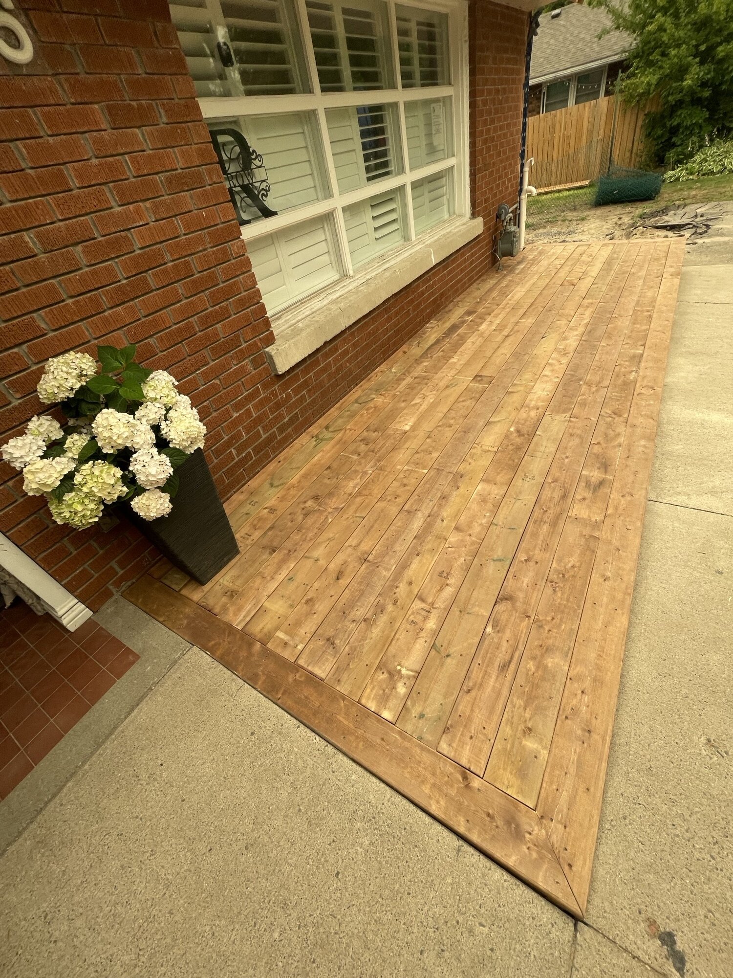 A new wood deck along a brick house, seen from the far end, with white hydrangeas in a planter by the window.