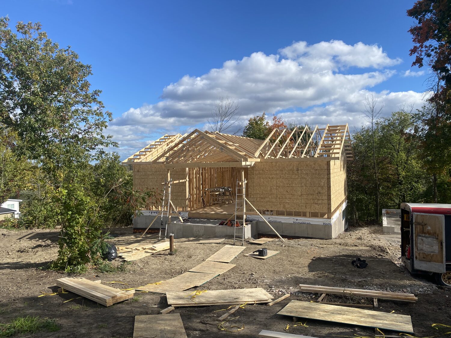 A new building framed on its concrete foundation, with roof trusses going up on a wooded lot.