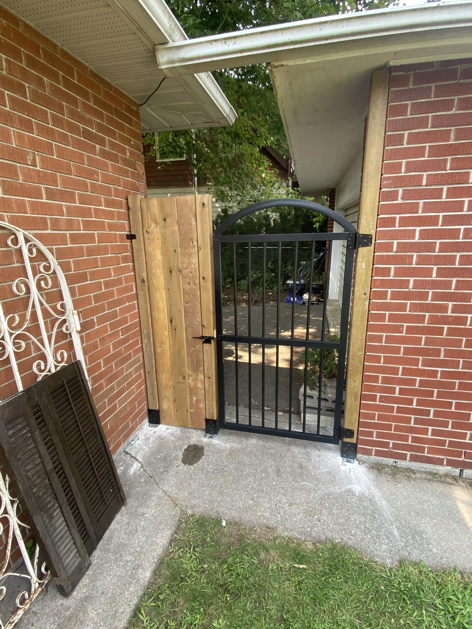A black arched metal gate between two brick walls, beside a new wood fence panel.