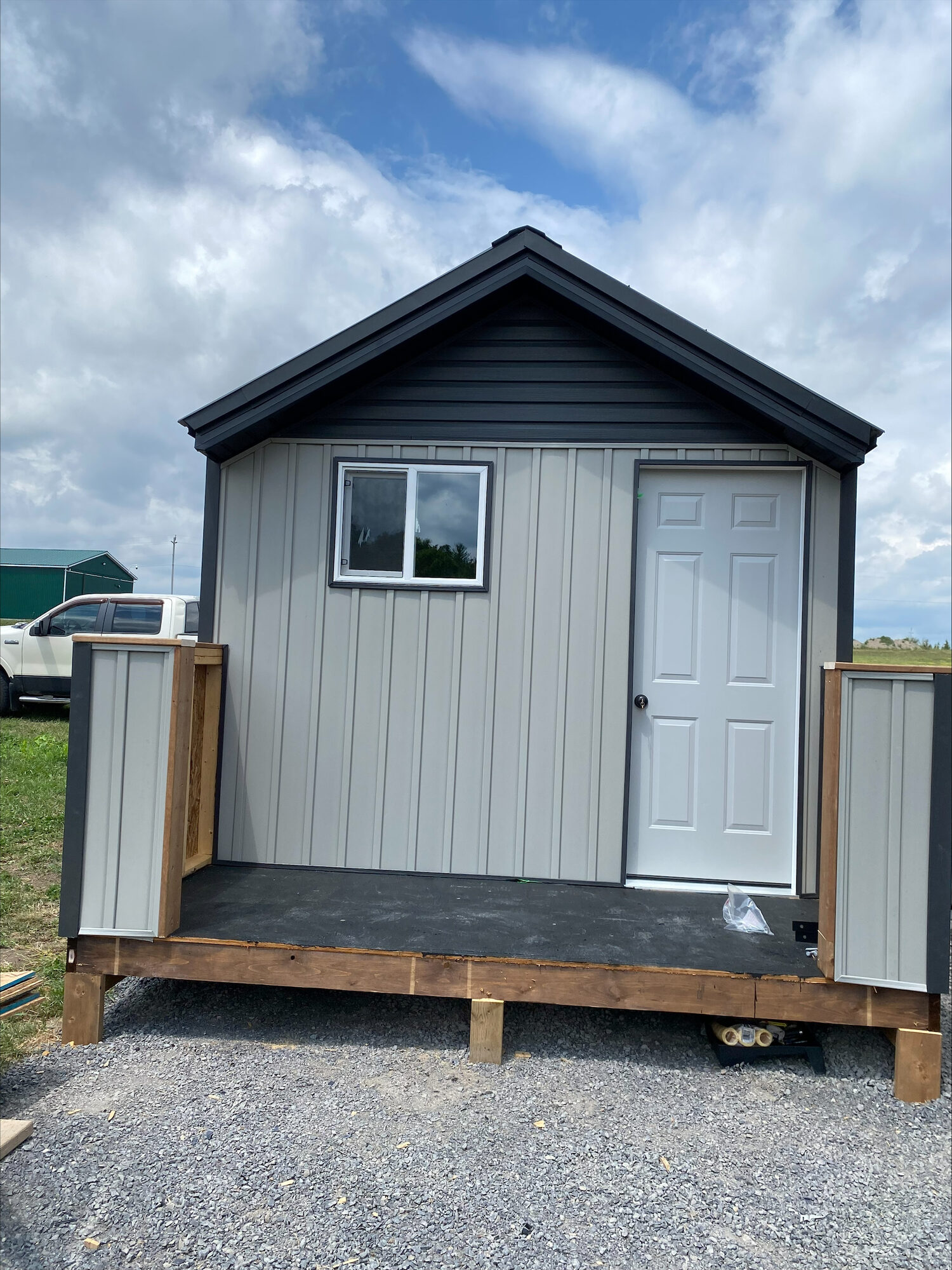 A new grey board and batten shed with a white door, a covered porch and a dark metal roof, set on timber posts over gravel.