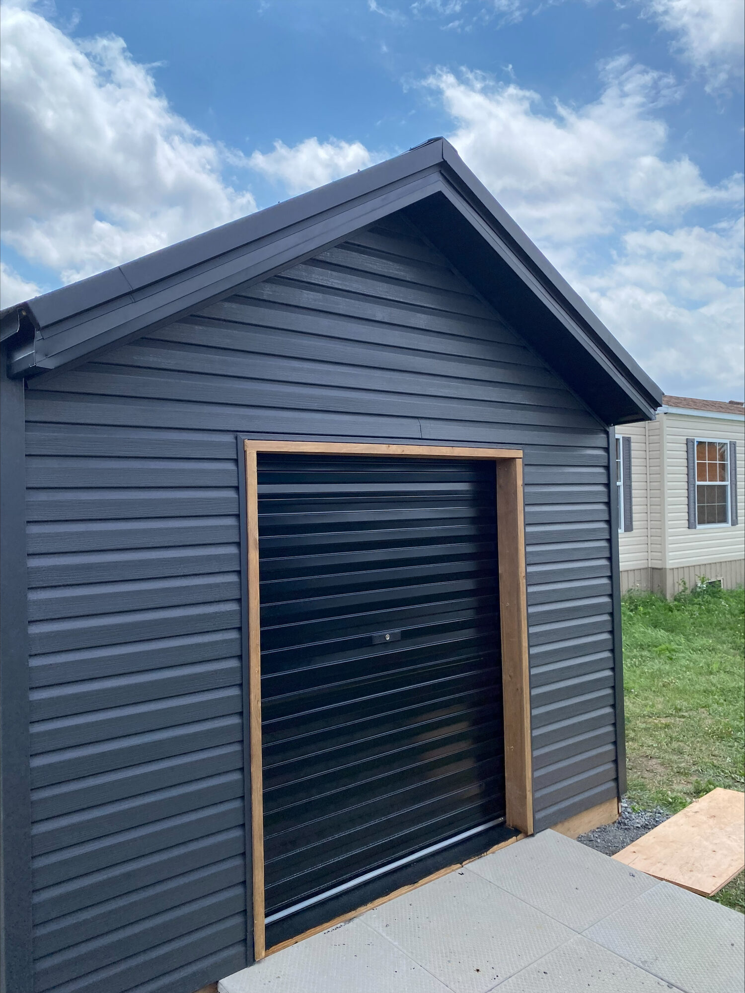 A navy sided outbuilding with a black roll up door, framed in natural wood trim.