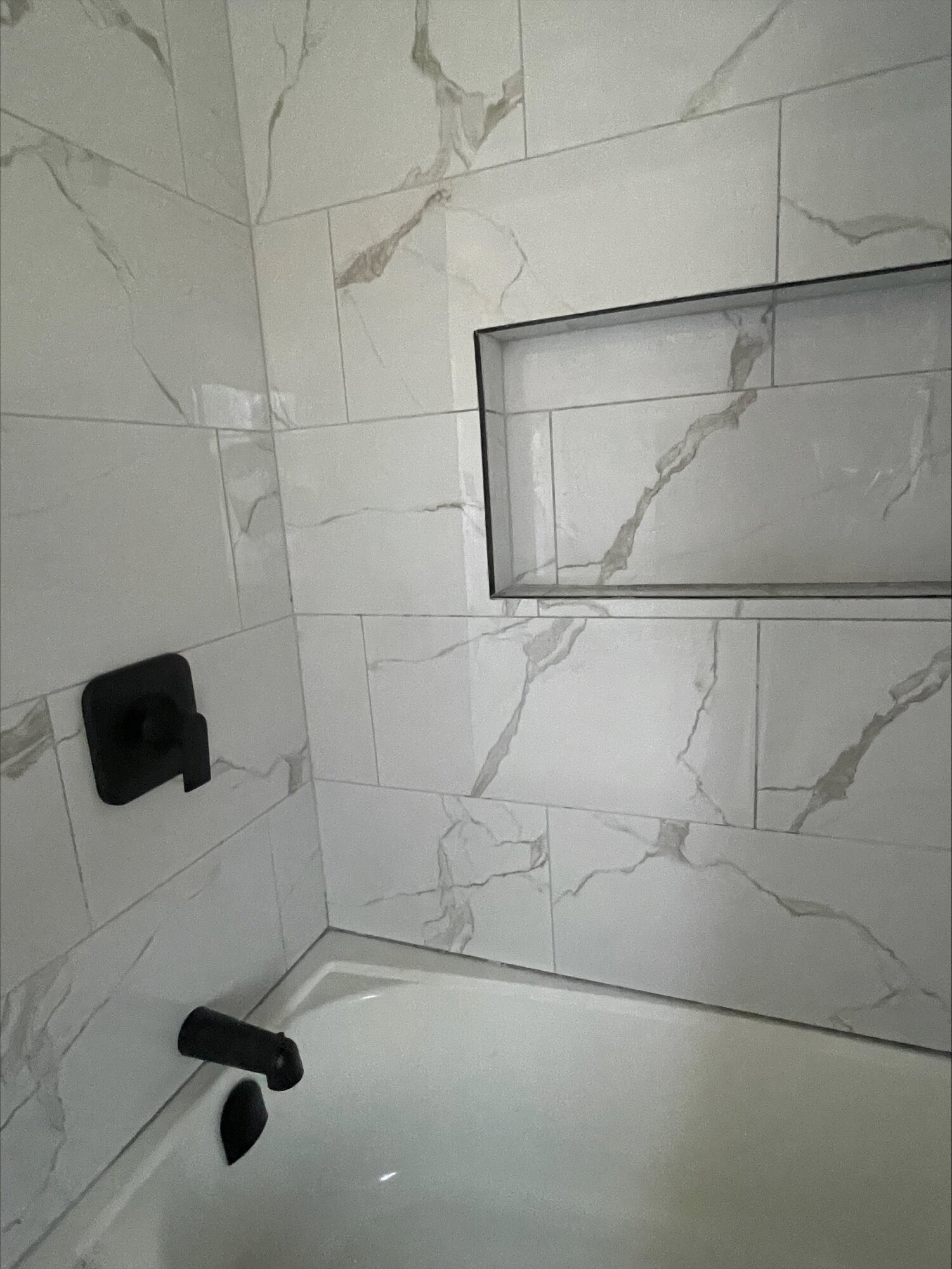 A marble look tile tub surround with black fixtures and a recessed niche above.