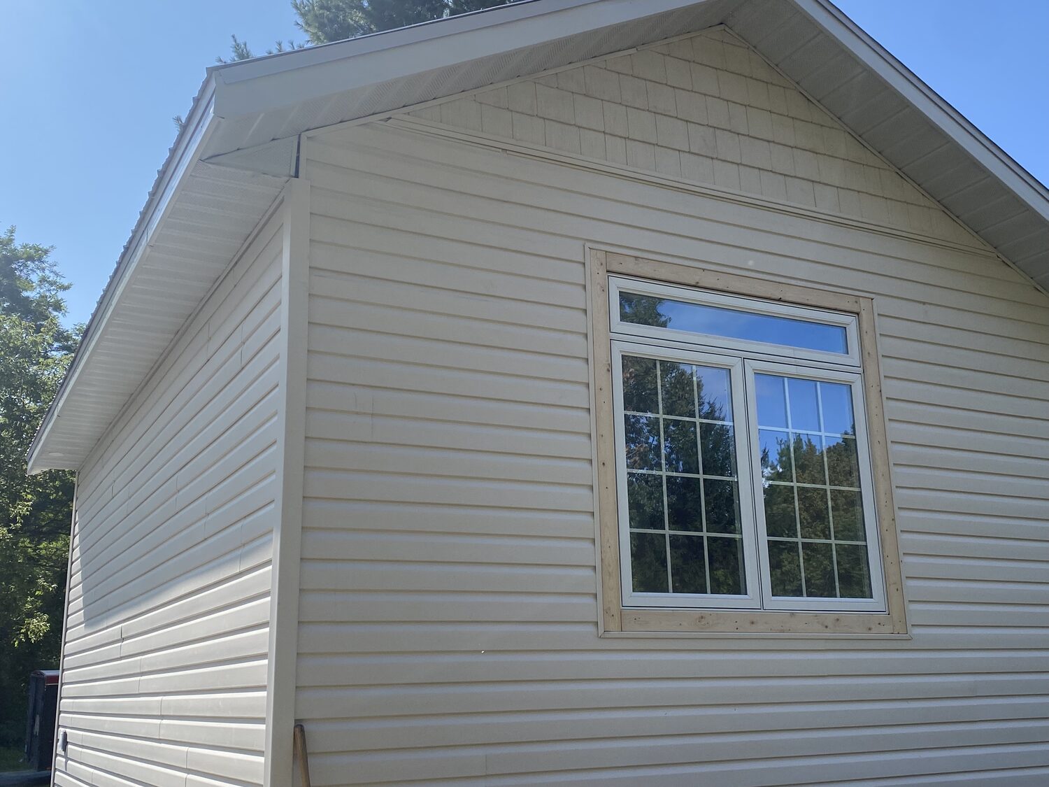 A new double window with a transom above, installed in a cream vinyl sided gable wall and framed in wood trim.